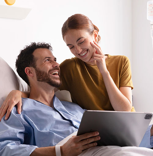 Mujer visitando a su pareja en el hospital, sonriendo y mirando una tablet