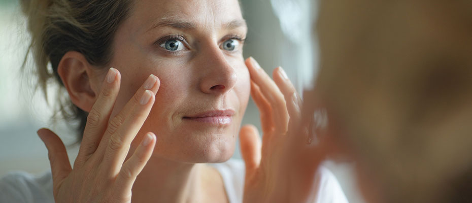 Mujer poniéndose crema facial frente a un espejo - Cuidados de la piel
