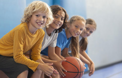 Niños y niñas sentados en un banco con una pelota de basketball en la mano | Deporte en la infancia: crecer fuertes, felices y saludables Niños y niñas sentados en un banco con una pelota de basketball en la mano | Deporte en la infancia: crecer fuertes, felices y saludables