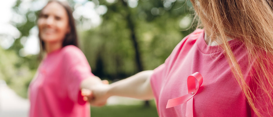 dos mujeres tomadas de la mano con los brazos estirados, de remera rosada y cinta rosada | Cáncer de mama: cómo cuidarte y detectarlo a tiempo