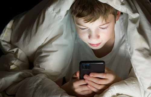 Adolescente mirando un celular debajo de un acolchado | Pornografía en niños, niñas y adolescentes