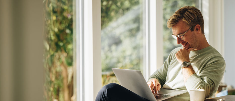 Hombre con un laptop en sus piernas, sentando en un comedor con una taza de café y sonriendo | Noviembre: información y prevención del cáncer de próstata