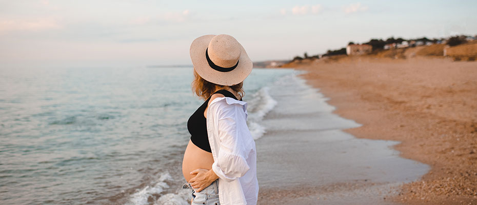 Mujer embarazada, de vaquero y camisa abierta en la orilla de una playa, mirando el hacia adelante | Embarazo en verano: ¿Cómo enfrentar el calor?