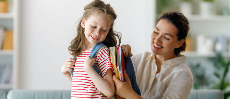 Madre guardando cuadernos en la mochila de su hija que la lleva en su espalda | Rutina y bienestar: cómo reencontrar el equilibrio