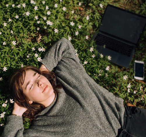 Mujer recostada en el pasto al sol con una computadora a su lado | Hacer una pausa también es cuidarte